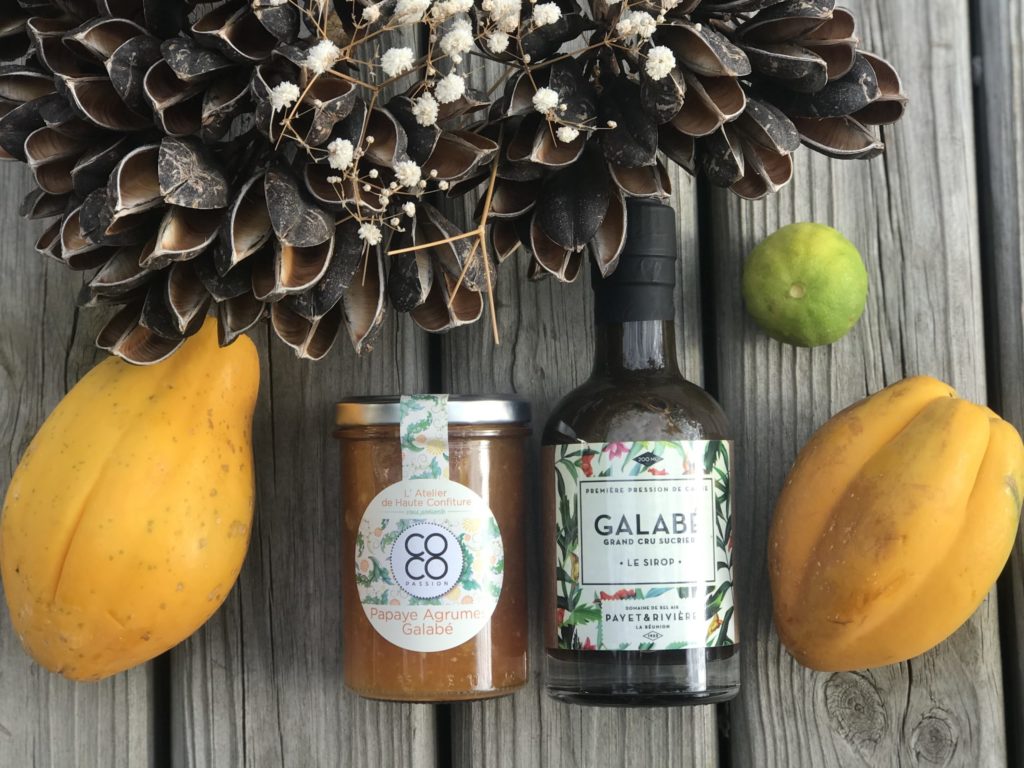 Un pot de confiture Coco Passion posé entre des papayes et une bouteille de galabé, sur du bois brut