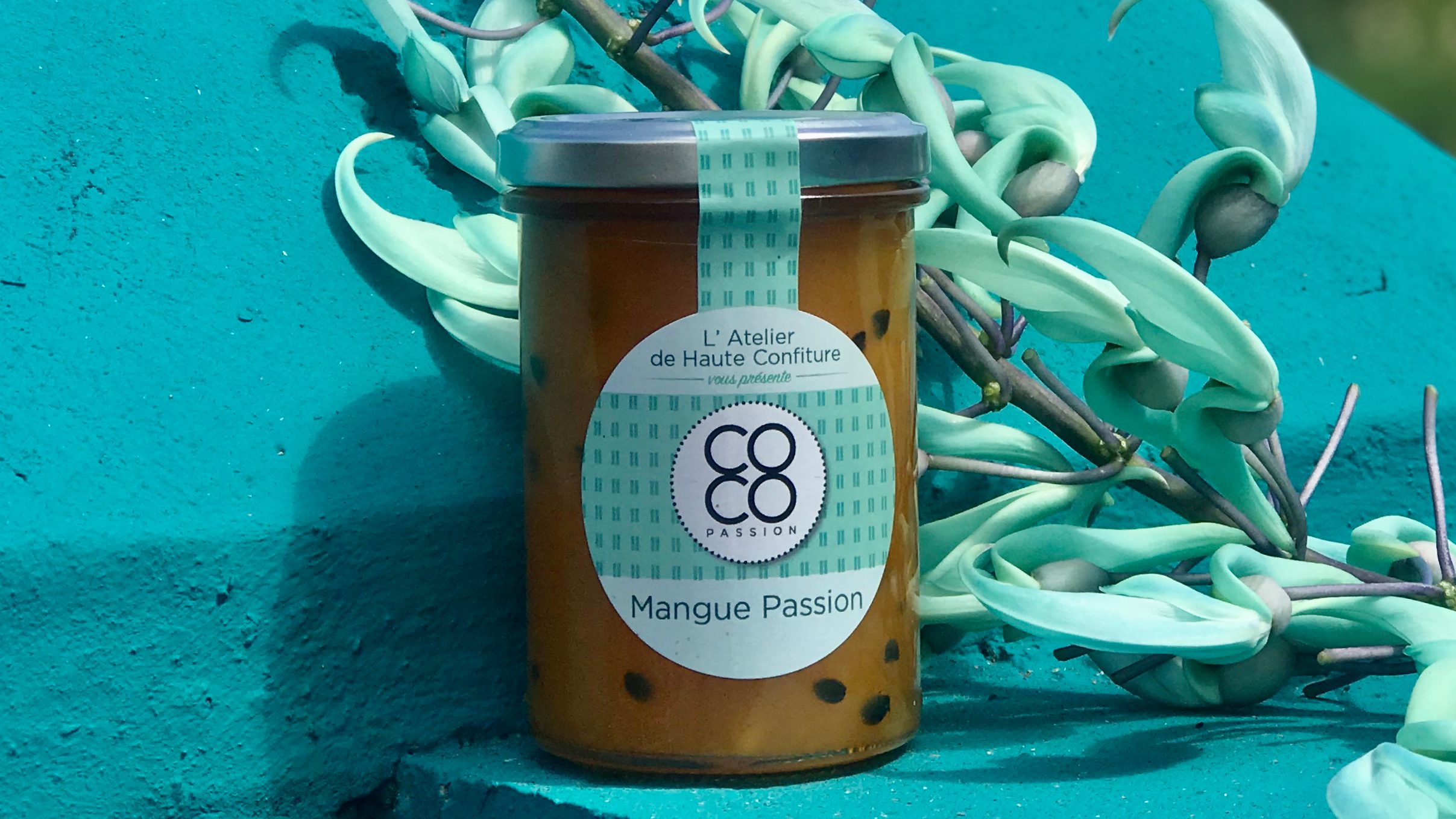 Un pot de confiture Mangue Passion posé devant un mur turquoise et des lianes