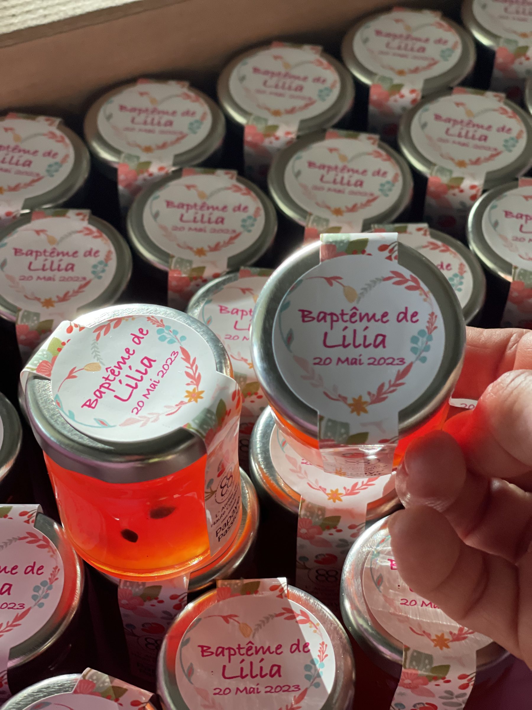 Petit pot de gelée tenu à la main, étiquette rose « Baptême de Lilia, 20 mai 2023 », devant d'autres pots identiques