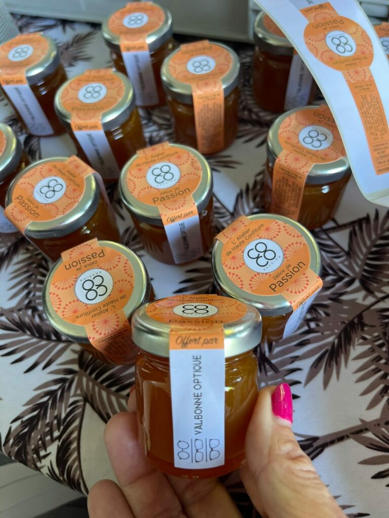 Petit pot de confiture passion « Offert par Valbonne Optique » tenu à la main, au-dessus d'autres pots