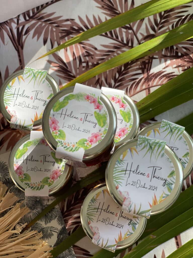 Petits pots de confiture aux étiquettes fleuries « Hélène & Thierry, 23 octobre 2024 », posés parmi des feuilles de palmier